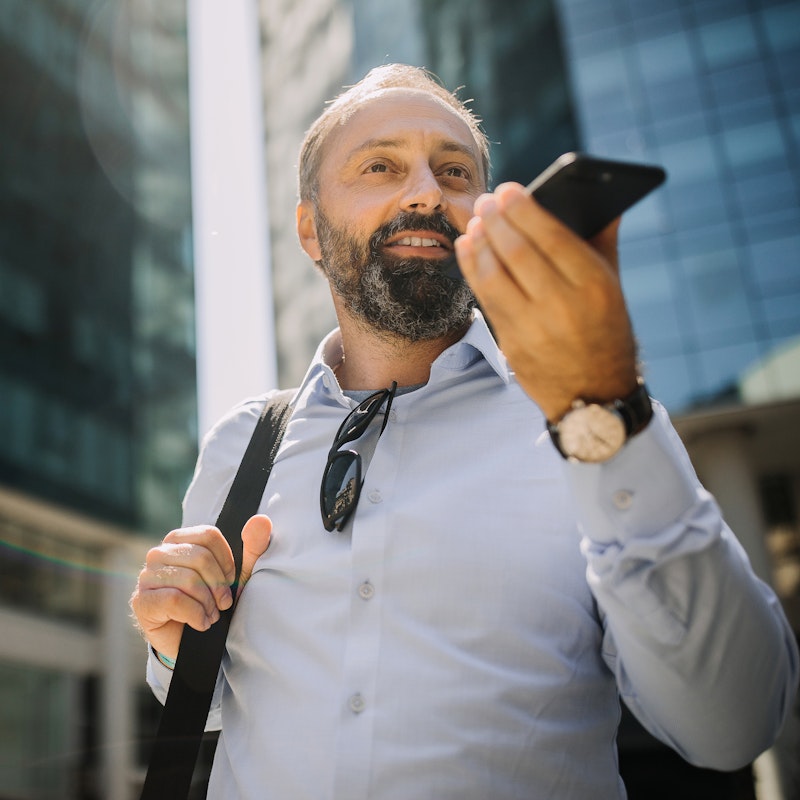 A man with a graying beard stands outside in front of a couple of mirrored skyscrapers. He wears a button-up shirt with a pair of sunglasses tucked into the collar and the strap of a bag over one shoulder. He holds a smartphone up near his mouth, ready to use it on speaker mode.
