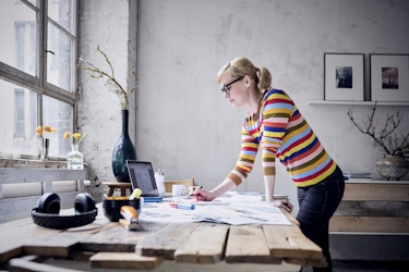  Woman working in her bright loft, standing over a desk of paperwork. 