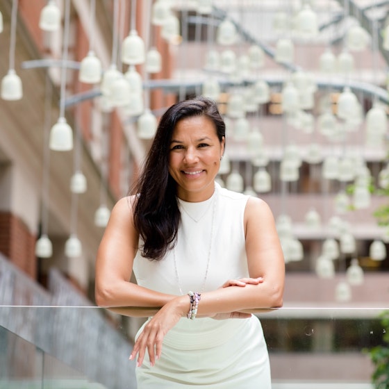 An image of Tiffany Houser, founder and CEO of EvolveEQ, leaning on a glass panel in the outdoor atrium of a multi-story building. Tiffany is a smiling woman with long, dark hair draped over one shoulder. She wears an ivory sleeveless top and a long, thin chain necklace.