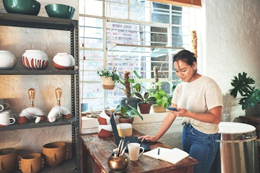  A woman stands next to a wooden table and looks at the smartphone in her hand. Her other hand rests on the keyboard of an open laptop on the table. On the other side of the table is a shelving unit filled with pottery. The table also holds some pots, including a small metallic gold pot filled with writing utensils. The windowsill next to the table is filled with potted plants. 