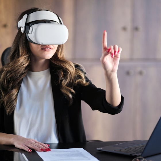 A woman wearing a VR headset sits at a desk and reaching up with one hand, a finger extended to poke or tap at something that only she can see. The table she sits at also holds an open laptop and a piece of paper covered with printed text.