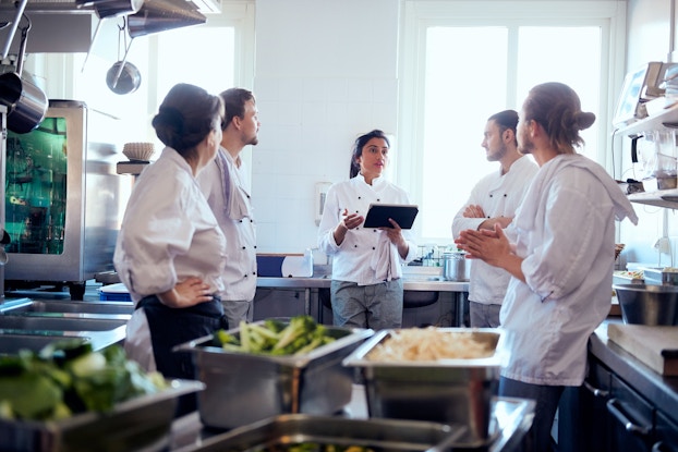  A group of five chefs in white jackets stand in a modern kitchen. The chef in the middle of the group, a woman with dark hair pulled back into a ponytail, is speaking while holding an electronic tablet. The other four chefs watch her. The kitchen is equipped with stainless steel equipment, including sinks, a few pots hanging from overhead vents, and a small glass-fronted refrigerator; in the foreground are a few stainless steel containers filled with vegetables.