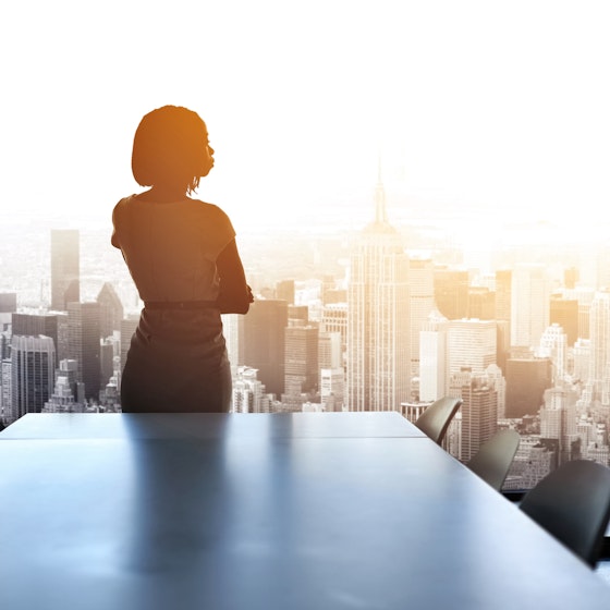 Professional woman standing in a conference room looking out on to the city through the floor-to-ceiling windows.